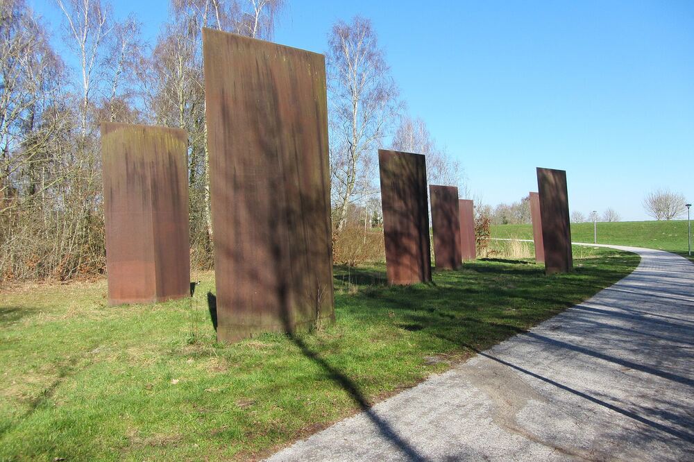 Rij hoge roestbruine stalen zuilen langs een wandelpad door het bos, met lange schaduwen op het gras