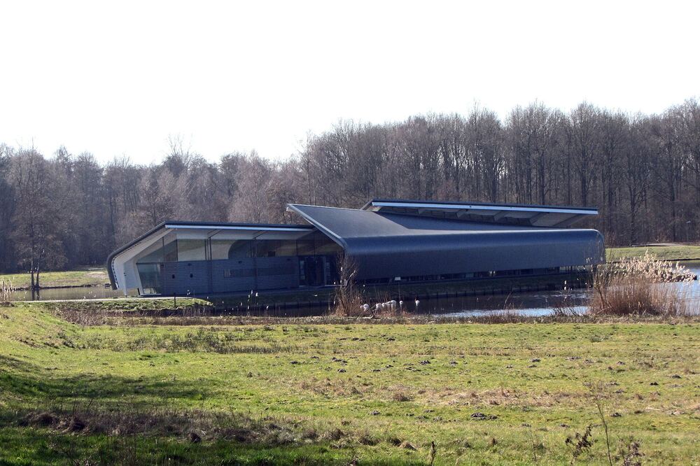 Laag donker paviljoen met schuin oplopend dak in een groene oever, met bos op de achtergrond